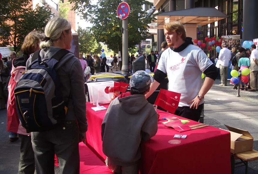 Drei Besucher*innen stehen an einem Infostand von on.tour und sprechen mit dem Mitarbeiter.