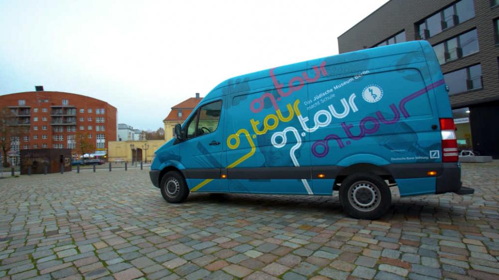 The blue on.tour bus is parked on a cobbled square. The words “on.tour” are written on the bus in pink, green, and blue. There are buildings in the background. 
