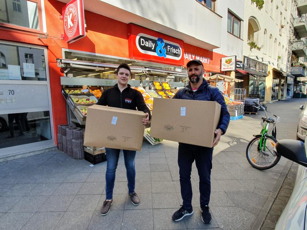 Two men stand in front of a grocery store with an orange outer wall and each hold a moving box in their hands.