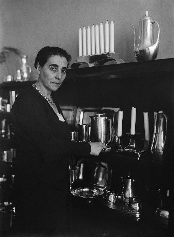 Emmy Roth stands in front of a shelf containing coffee and tea sets and candlesticks