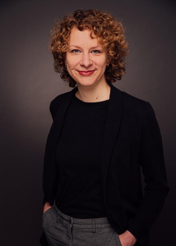A woman with red curly hair and black blazer stands in front of a gray background and looks into the camera.