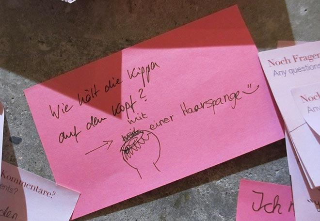 Post-it note in pink: "Wie hält die Kippa auf dem Kopf? - mit einer Haarspange" (How does a kippah stay on? - with a hair clip))