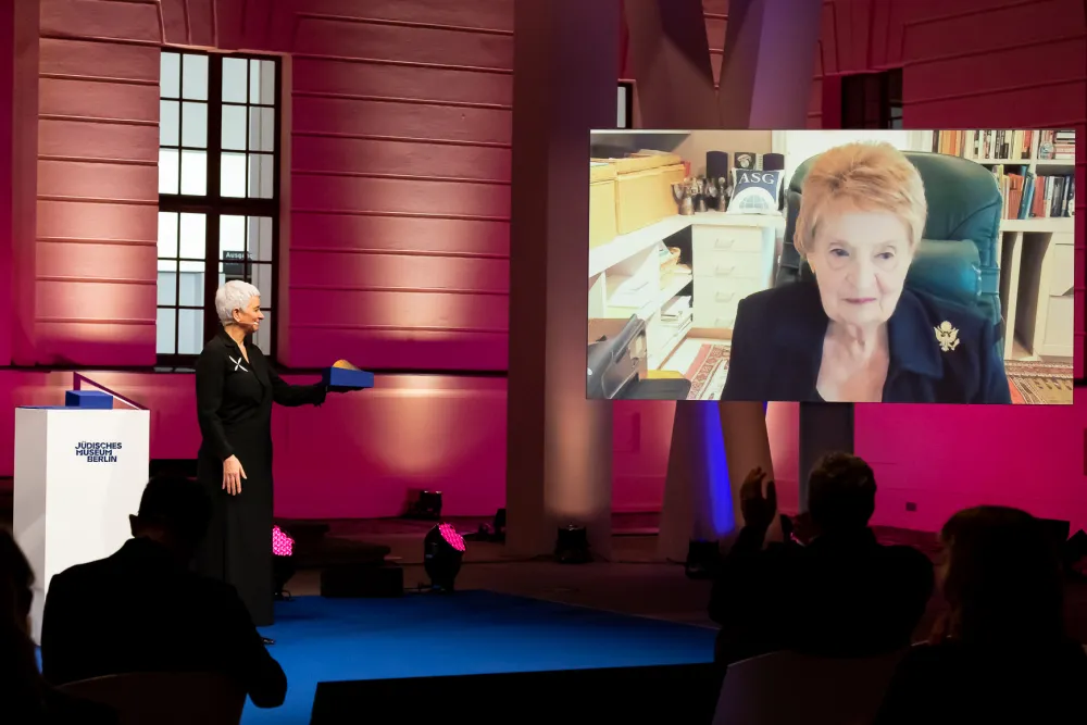 Hetty Berg stands on a stage, Madeleine Albright is on a screen via video call