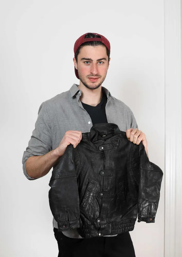 A young man with a red cap holds a black leather jacket up to the camera.