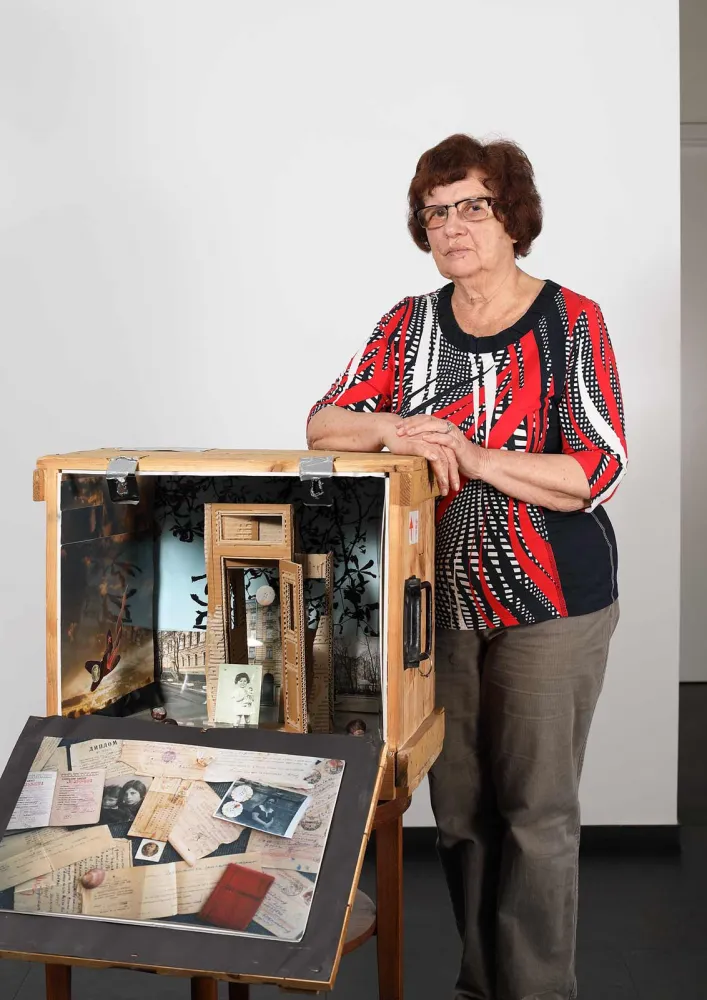 An older woman with reddish hair stands next to an open wooden box, which is designed like a stage with various objects.