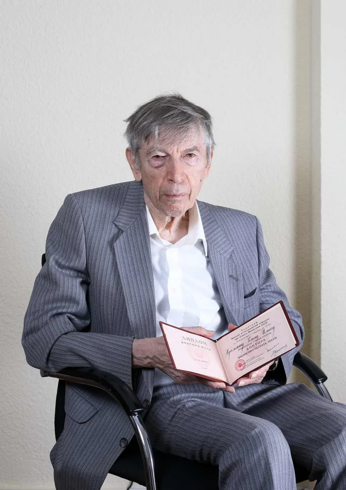 An older gentleman in a gray-striped suit sits on a chair and holds a certificate up to the camera.