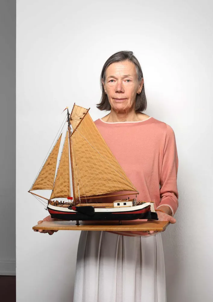 A woman with graying hair, an apricot sweater and a white skirt holds a wooden board with a model ship on it up to the camera.