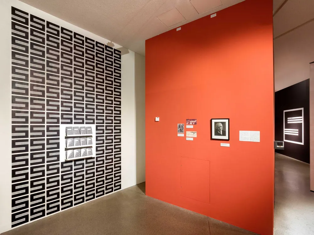 View into an exhibition room, with a central orange-colored wall on which hangs a framed portrait photo of Salman Schocken, on the wall to the left the S-shaped Schocken logo in black and white several times as well as some brochures.