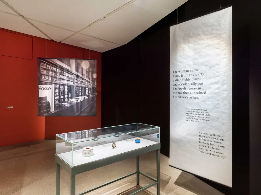 View into an exhibition room with a display case showing a teapot, a tin can and a porcelain can, with a close-up of a department store in the background.