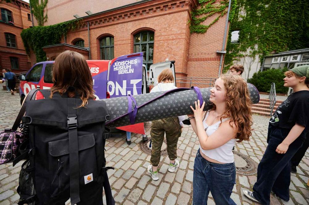 Young people carry a rolled-up carpet in a paved courtyard.