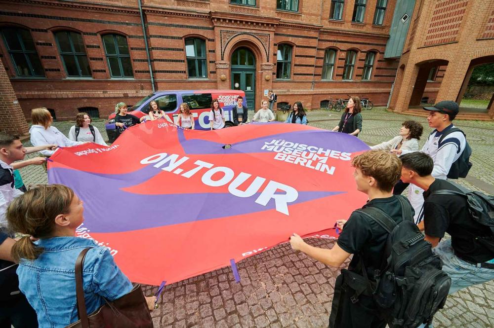 Eine Gruppe von Jugendlichen spannen auf einem Innenhof ein großes Tuch auf, auf dem on.tour und Jüdisches Museum Berlin steht.