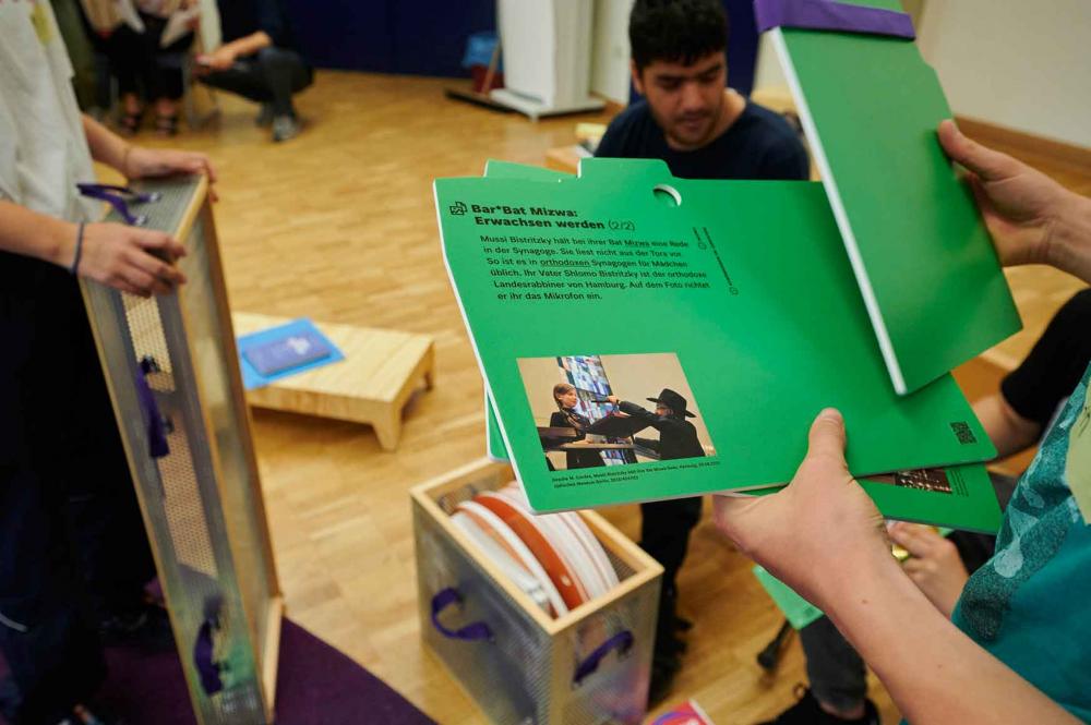 Hands are holding green cards with a photograph and text on them, titled in German Bar*Bat Mitzvah: Growing Up.