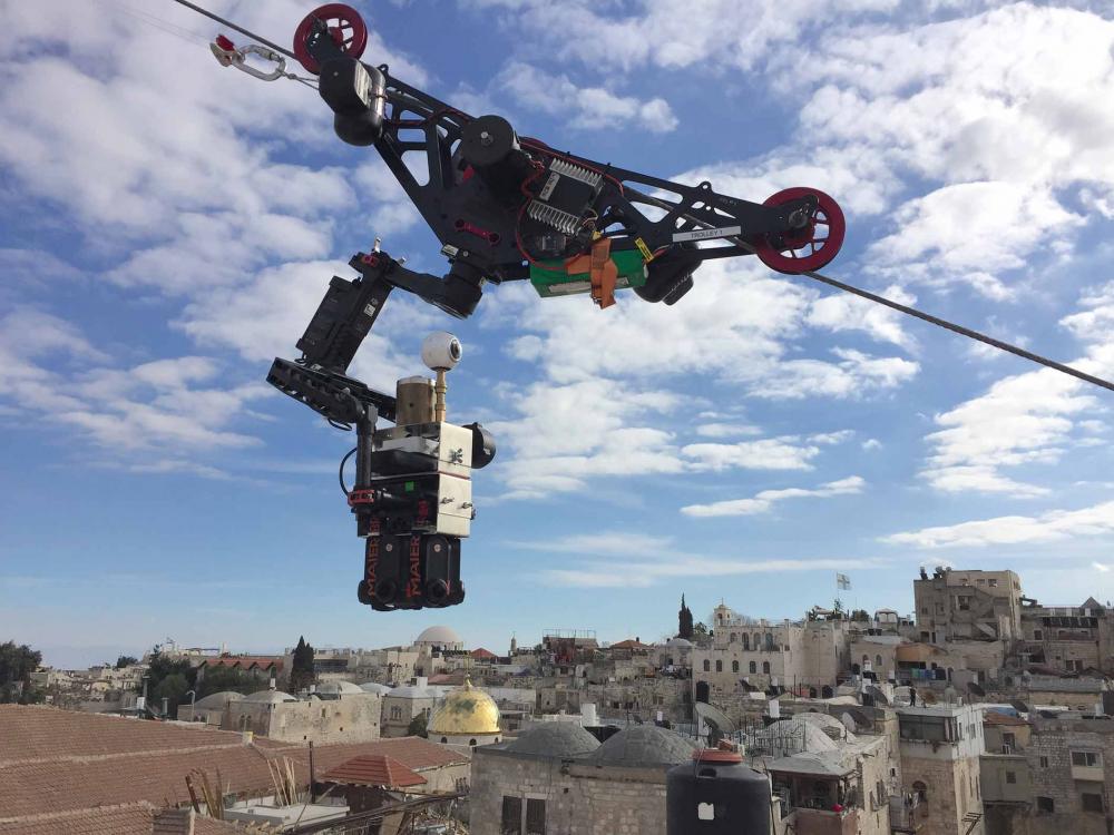 A camera mounted on a steel cable hovers before a blue sky over the rooftops of Jerusalem.