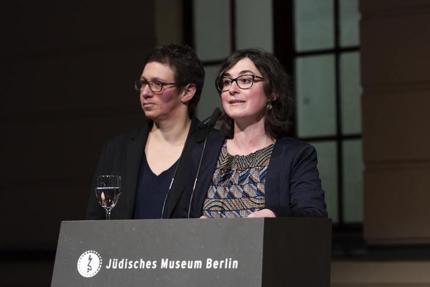 Alina Gromova is standing at a desk on which the logo and lettering of the Jewish Museum are written. There is a glass of water on the desk. Susan Kamel is standing behind her.
