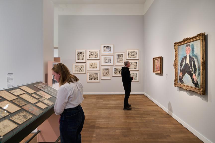 Exhibition space with visitors and paintings on white wall, a showcase with paintings and drawings in the center of the room.