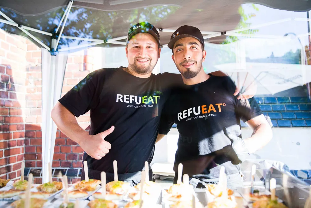 Two men smile into the camera, a table with food in front of them.
