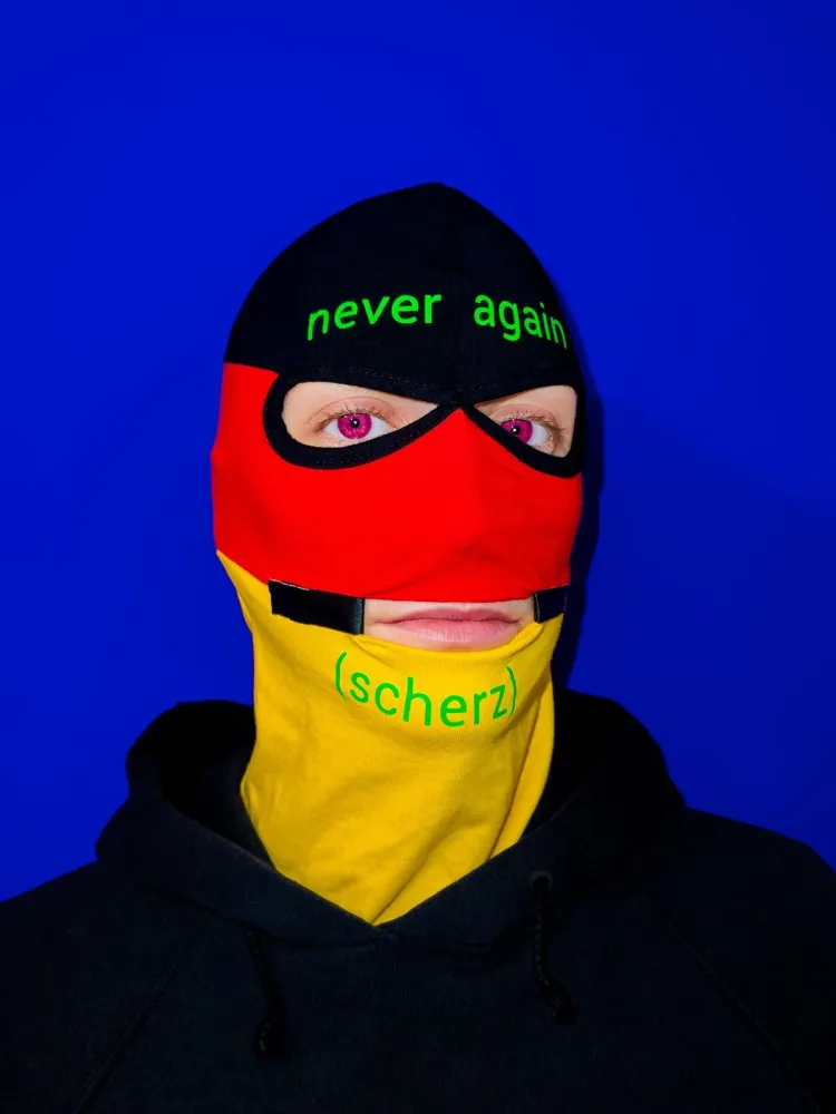 Portait Foto einer maskierten Person vor blauem Hintergrund. Die Person tr&auml;gt eine schwarz-rot-gelbe Sturmhaube mit der Aufschrift never again auf der Stirn sowie (scherz) auf dem Kinn.