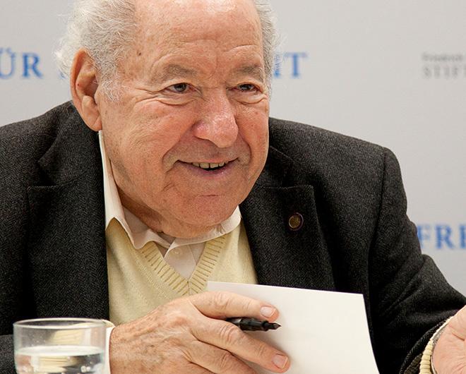 Photography: A smiling elderly man (Sally Perel) holds a pen in his hand and sits in front of an open book.