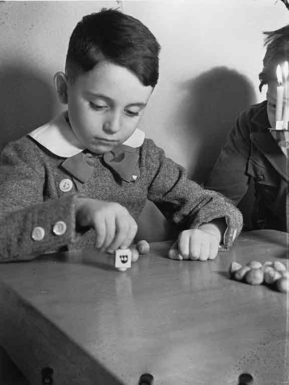 Schwarz-Weiß-Fotografie, die einen Jungen mit Dreidel an einem Tisch zeigt. Am Bildrand ist ein zweiter Junge zu sehen