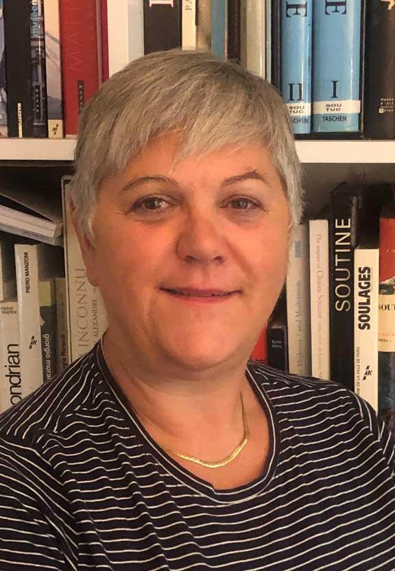 A woman with short gray hair stands in front of a bookshelf and looks into the camera. 