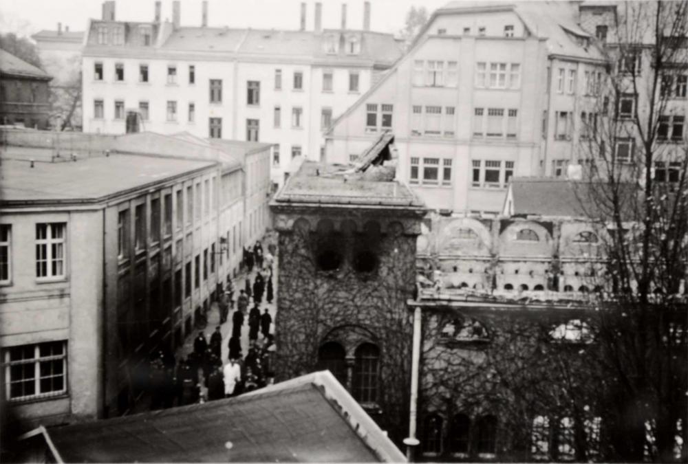 Das Fotot zeigt eine zerstörte Synagoge, deren Dach fehlt, es scheint verbrannt zu sein, Trümmer liegen auf dem noch bestehenden Eingangsportal, in der Staße davor befinden sich viele Passanten.  