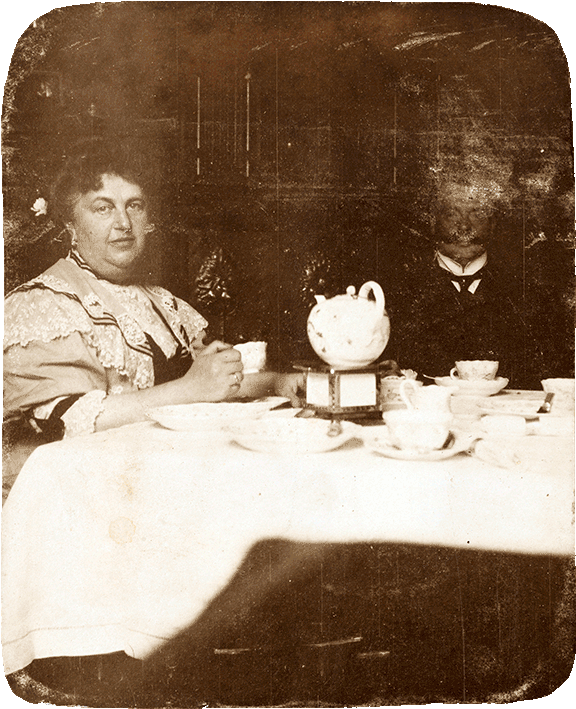 Heavily weathered black-and-white photo: It shows a middle-aged couple sitting at a table set for coffee. Both are looking toward the camera.
