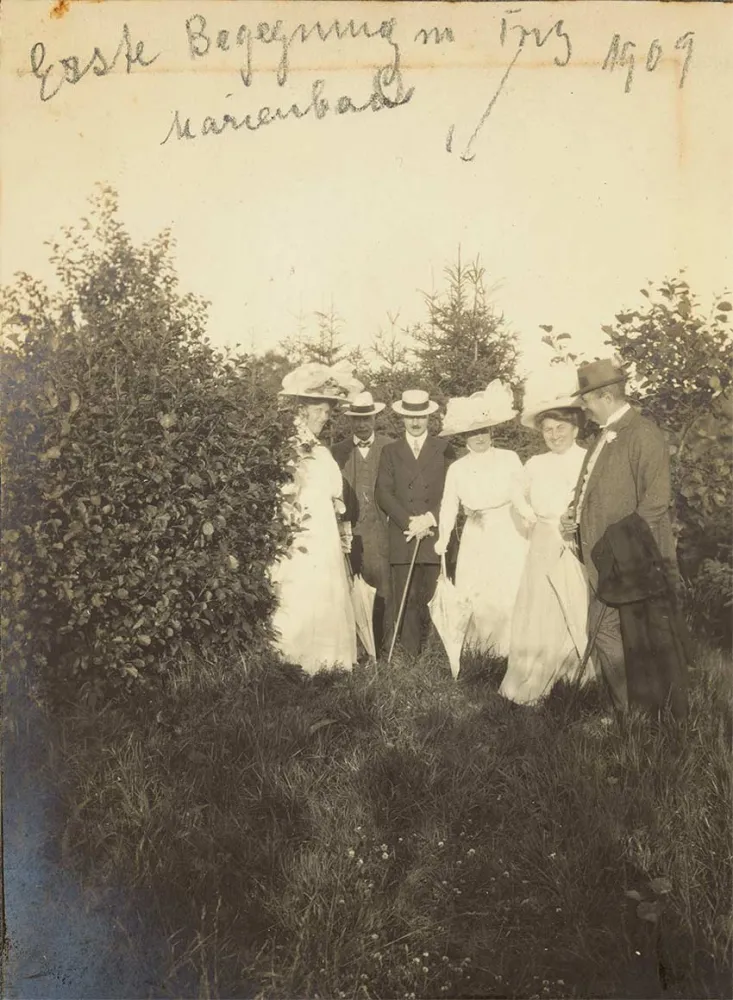 Drei Herren in Anzug mit Hut und Stock und drei Damen in weißen Kleidern mit großen Hüten und Sonnenschirmen auf einer Lichtung. Oben auf dem Foto steht in Handschrift: „Erste Begegnung m Fritz Marienbad 1909“, ein Pfeil weist auf Fritz.
