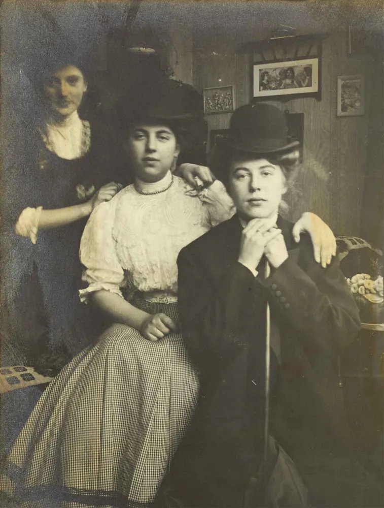Schwarz-Weiß-Foto von drei jungen Frauen, sie haben sich schräg hintereinander positioniert, die vorderen beiden Damen sitzen, die hintere steht. Im Hintergrund ist eine mit Bildern behängte Wand zu sehen.