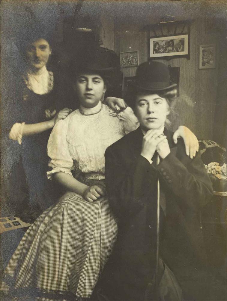 Schwarz-Weiß-Foto von drei jungen Frauen, sie haben sich schräg hintereinander positioniert, die vorderen beiden Damen sitzen, die hintere steht. Im Hintergrund ist eine mit Bildern behängte Wand zu sehen.