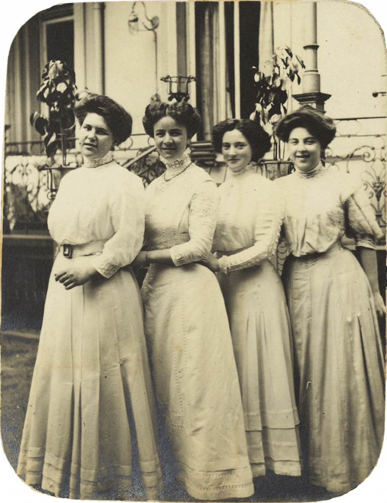 Schwarz-Weiß-Foto, das vier in einer Reihe schräg hintereinander stehende junge Damen im Dreiviertelprofil vor einem Haus zeigt. Alle tragen weiße, hochgeschlossene Kleider und Hochsteckfrisuren.