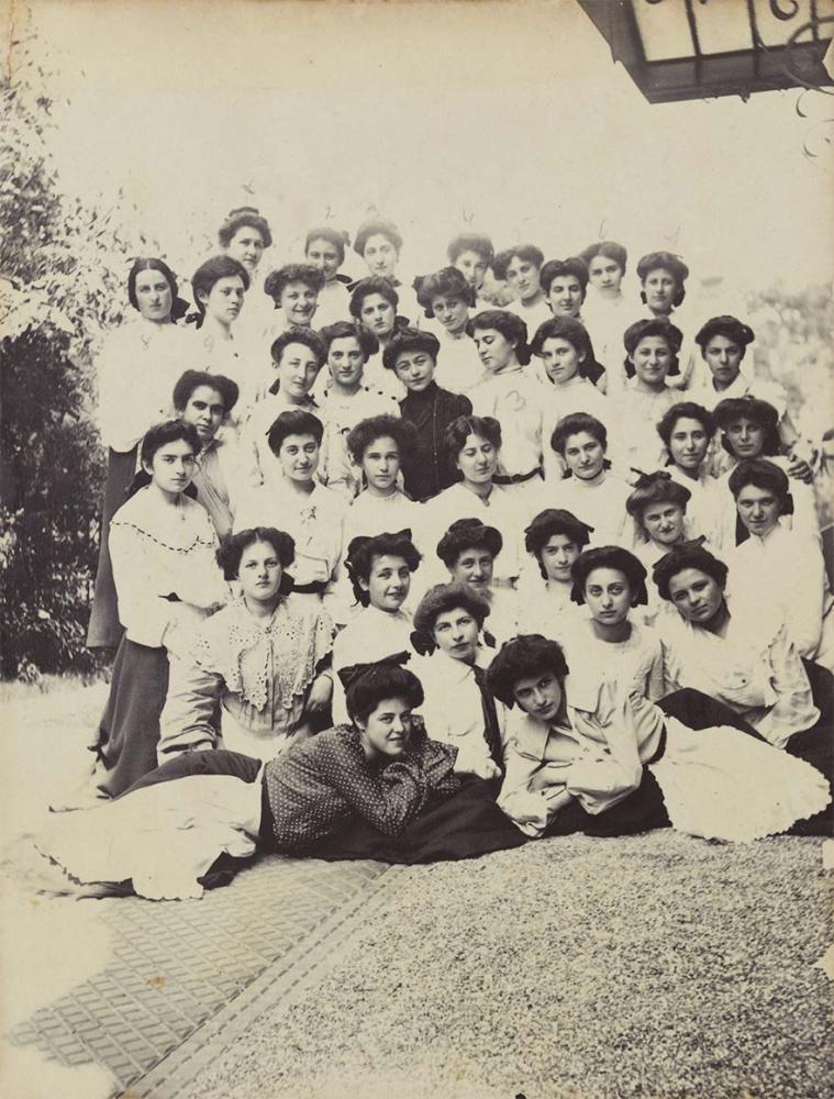 Black and white photo of a group of about 40 young women outdoors, they are next to a building that is visible slightly cropped at the right edge of the picture. Almost all of them are wearing updos, white blouses and dark skirts with white aprons.