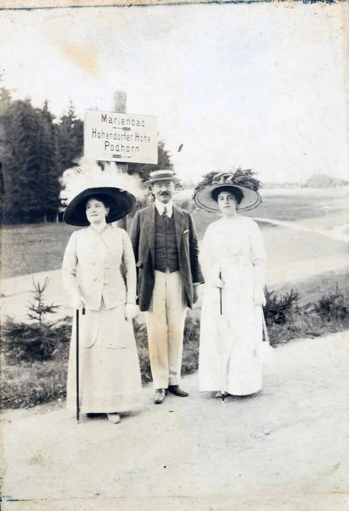 Das Foto zeigt einen Herrn zwischen zwei Damen auf einem Spaziergang. Sie posieren vor einem Wegweiser, der für „Marienbad“ nach links und für „Hohendorfer Höhe“ und „Podhorn“ nach rechts weist.