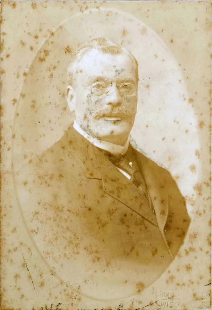 Stockfleckiges Porträtfoto eines gesetzten Herren im Anzug. Er trägt eine Krawattennadel, Schnurrbart und Monokel und wirkt sehr distinguiert.