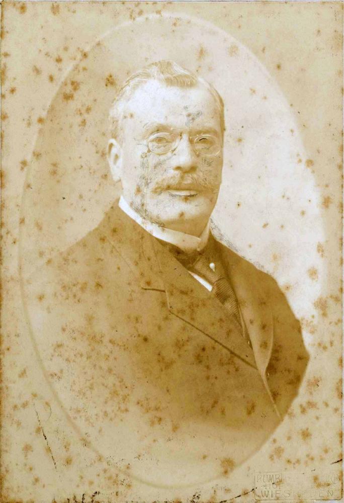 Portrait photo with foxing showing a staid gentleman in a suit. He wears a tie pin, mustache and monocle and looks very distinguished.
