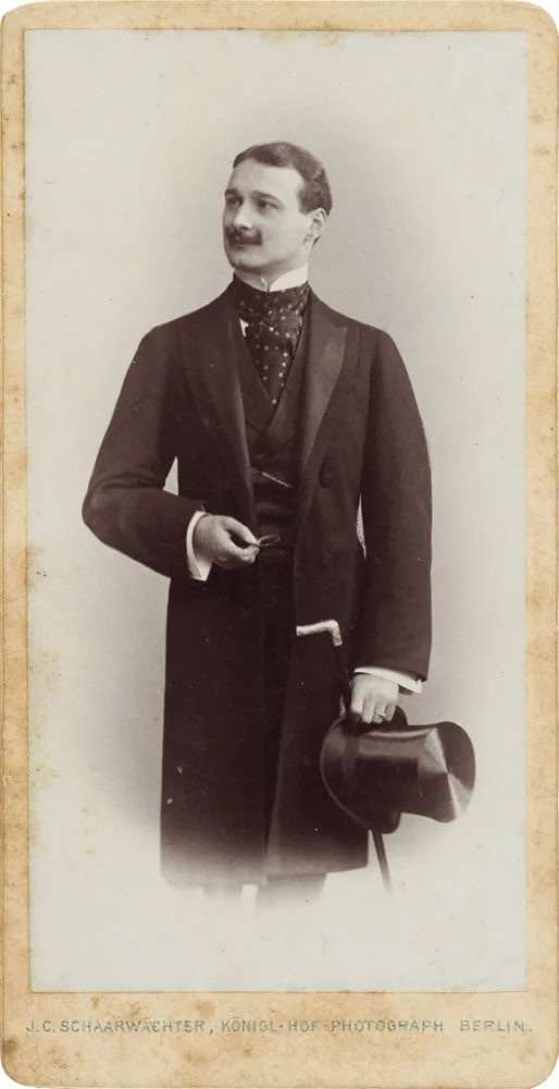 Schwarz-Weiß-Porträtfoto eines stehenden jungen Mannes. Der Körper ist frontal, der Kopf nach links gedreht im Dreiviertelprofil zu sehen. Er trägt einen Schnurrbart und einen Gehrock, in der Hand hält er einen Zylinder und Gehstock mit Fritzgriff.