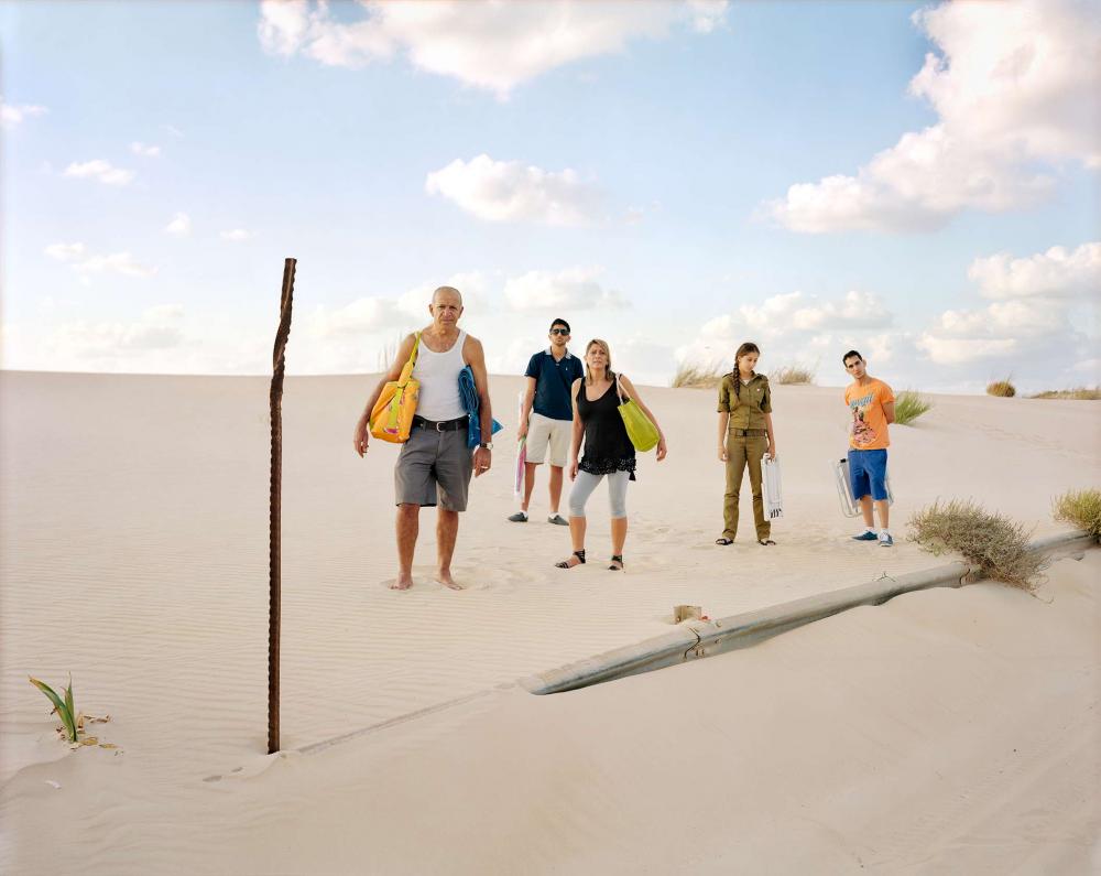 Eine fünfköpfige Familie steht im Sand auf einer Düne mit Strandtaschen und Klappstühlen, die Tochter in Armeekleidung
