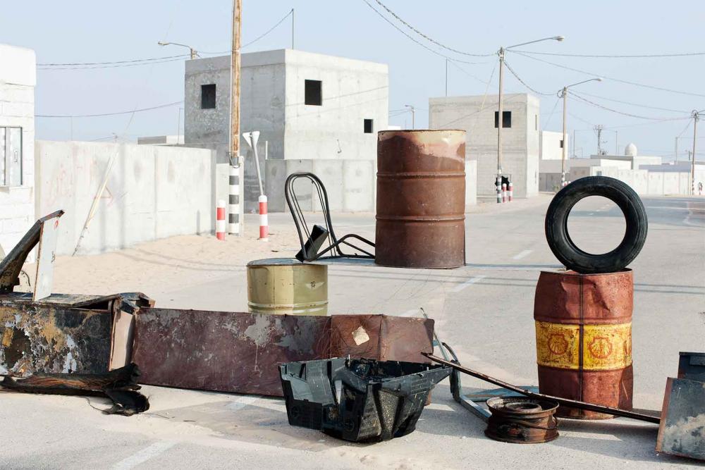 Tin barrels and other sheet metal objects and a car tire on a road along white flat-roofed houses