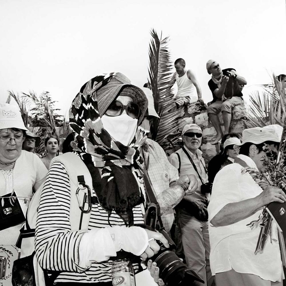 Schwarz-weiß-Foto eines Menschenzugs mit Palmzweigen, im Vordergrund eine Frau mit Fotoapparat, Hut, Kopftuch, Sonnenbrille und Mundschutz