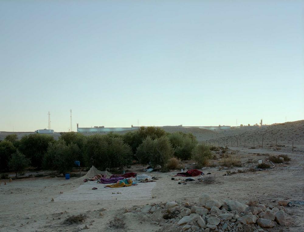 Outdoor sleeping camp on sandy and stony ground, behind it a few trees, on the horizon buildings in the morning light