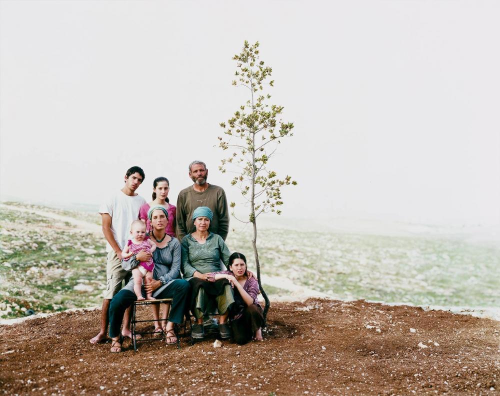 Sieben Menschen verschiedenen Alters, darunter ein Kleinkind, in der Pose eines Familienfotos im Freien neben einem jungen Bäumchen vor sonst kahler Landschaft