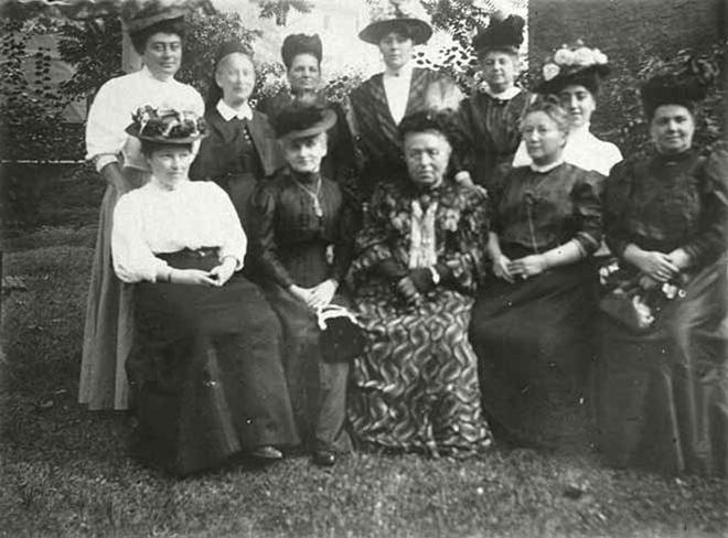 Gruppenbild von elf Frauen mit ausgefallenen Hüten in einem Garten