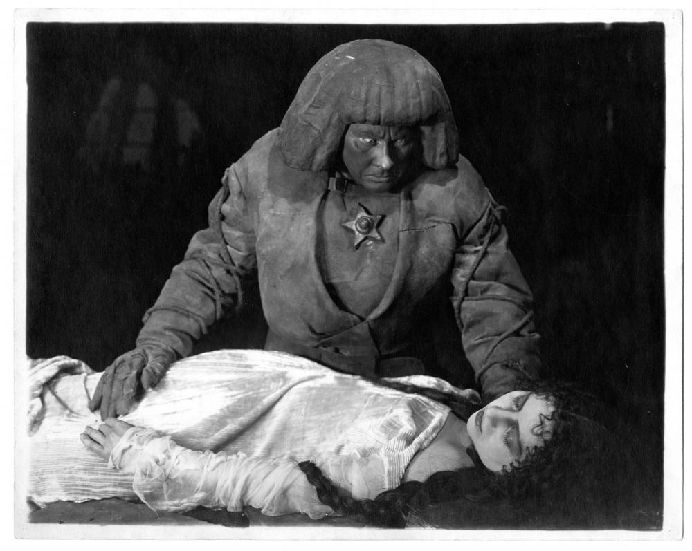 Filmstill aus dem Film„Der Golem, wie er in die Welt kam“ von Paul Wegener: Der Golem blickt erschrocken auf eine liegende Frau, seine Hand auf ihrem Bauch.