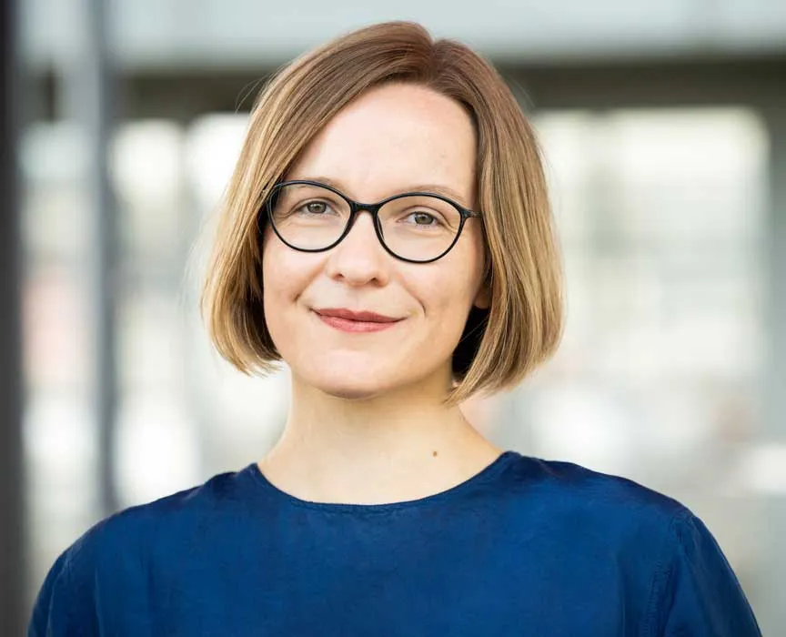 A woman with short blond hair, black glasses and a blue sweater smiles at the camera. 