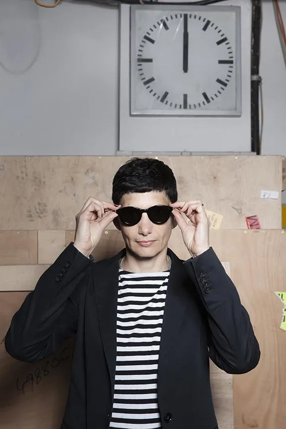 Porträt (Brustbild) einer Frau mit schwarzem kurzem Haar. Sie trägt ein schwarzes Jackett und ein schwarz-weiß quergestreiftes T-Shirt und setzt sich gerade eine dunkle Sonnenbrille auf. Im Hintergrund ist eine Wanduhr zu sehen, die 12 Uhr anzeigt.