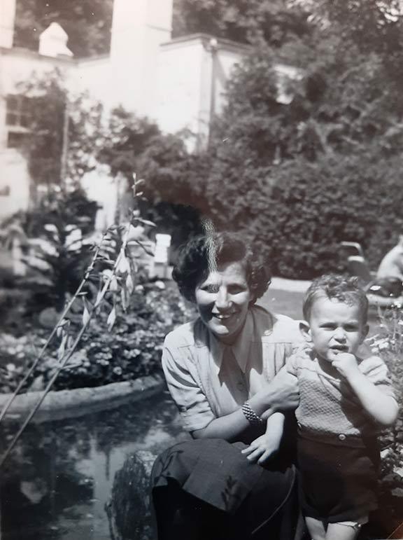Black and white photo of a toddler in leather pants, next to him kneels a woman in skirt and blouse, in the background a house and many plants.