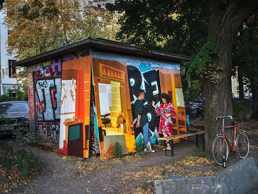 A man in jeans, lumberjack shirt and sneakers and a barefoot woman in a colorful dress are facing each other in front of a brightly painted shed, in the front right a bicycle leans against a tree, on the ground in front of it is a beer bottle