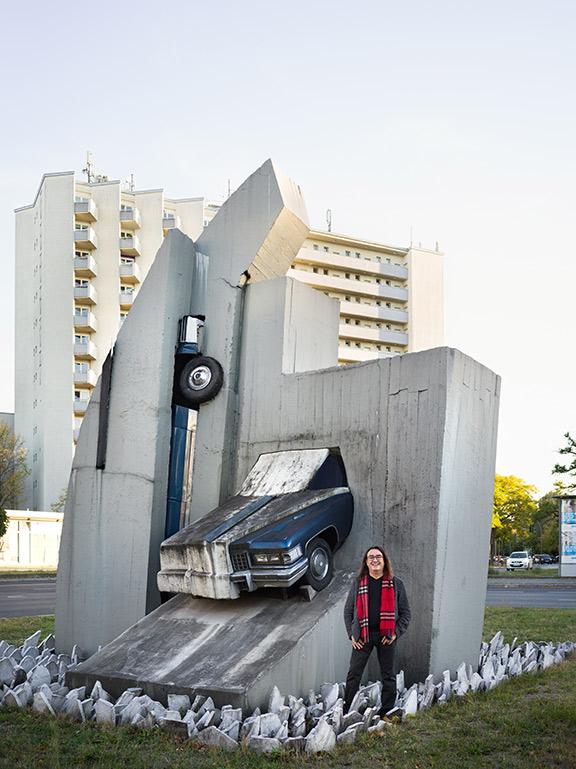 A man dressed in gray and black with a red scarf stands in front of a sculpture with half a car emerging diagonally from a concrete wall, in the background a tall apartment block