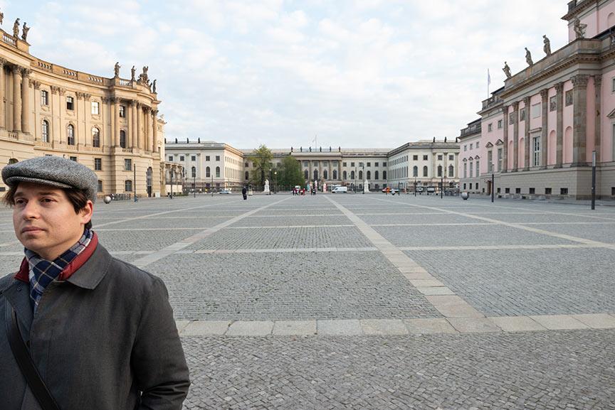 Panorama-Ansicht des Bebelplatzes in Berlin, rechts die Staatsoper, im Hintergrund die Humboldt-Universität, vorne links im Bild ein junger Mann mit Schirmmütze und Mantel