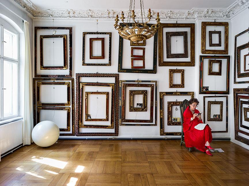 A woman dressed all in red sits on a chair in a room with many empty frames on the wall and a chandelier on the ceiling and looks at her cell phone