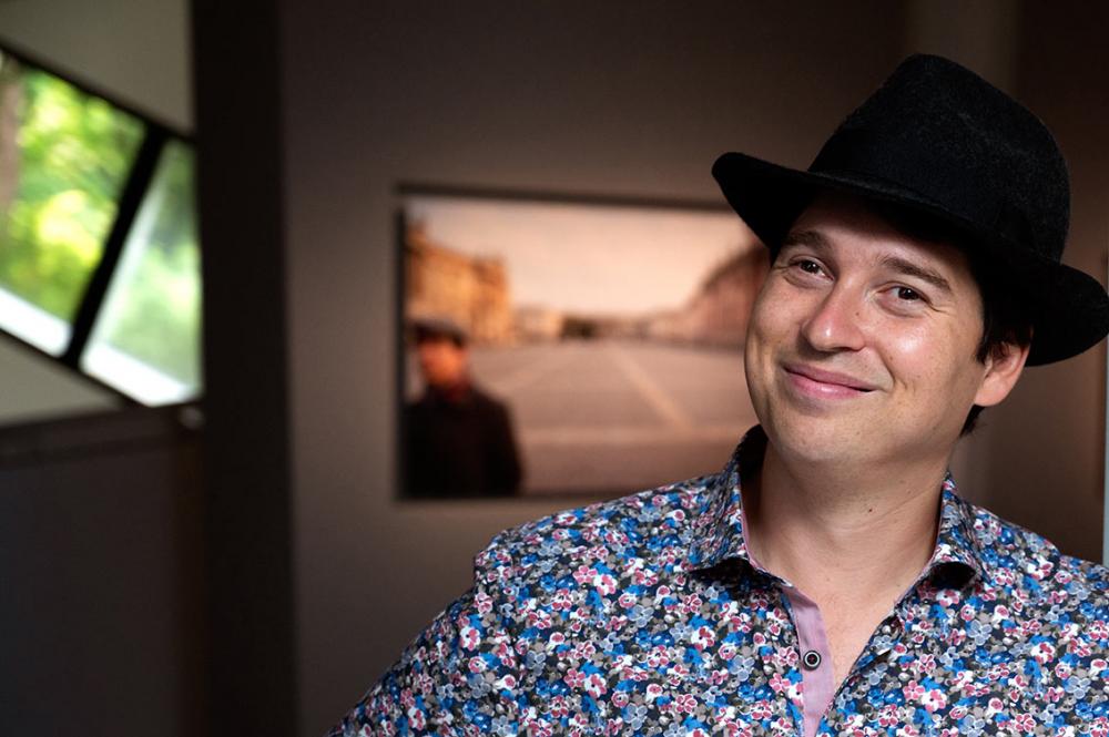 A man in a black hat and floral shirt laughs into the camera, in the background a portrait of him blurred on an exhibition wall